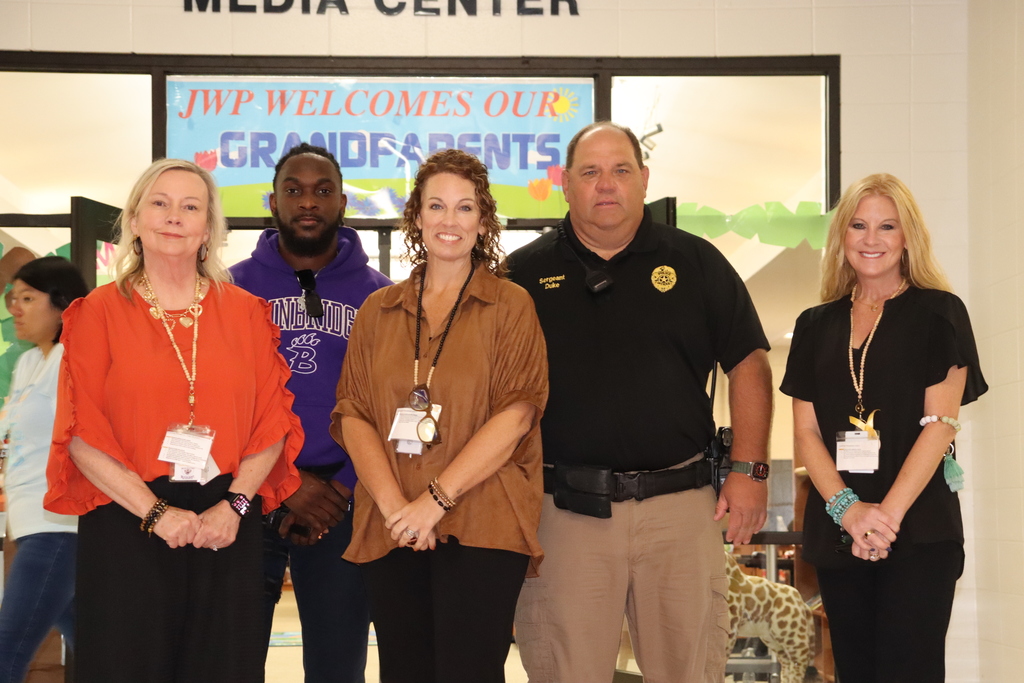 Five staff members and a school resource officer stand in front of the media center doors under a colorful banner that reads, "JWP Welcomes Our Grandparents."