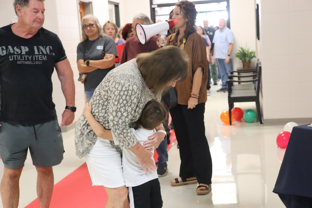 A woman crouches down to hug a young boy in a blue shirt while other adults and children walk by in a crowded school hallway.