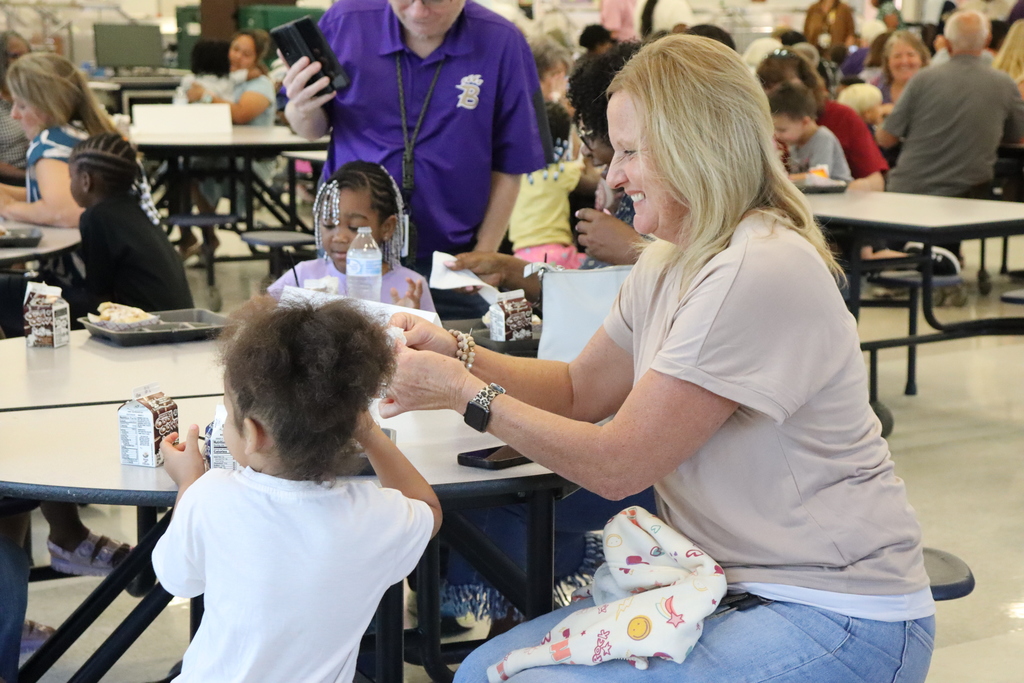 A woman sits at a lunchroom table smiling as she helps a young girl with her milk carton while other families eat in the background.