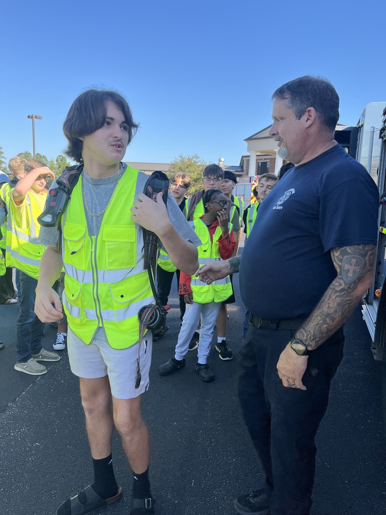 A student wearing a safety vest tries on a firefighter’s breathing apparatus while a BPS firefighter instructs him, as classmates watch in the background.