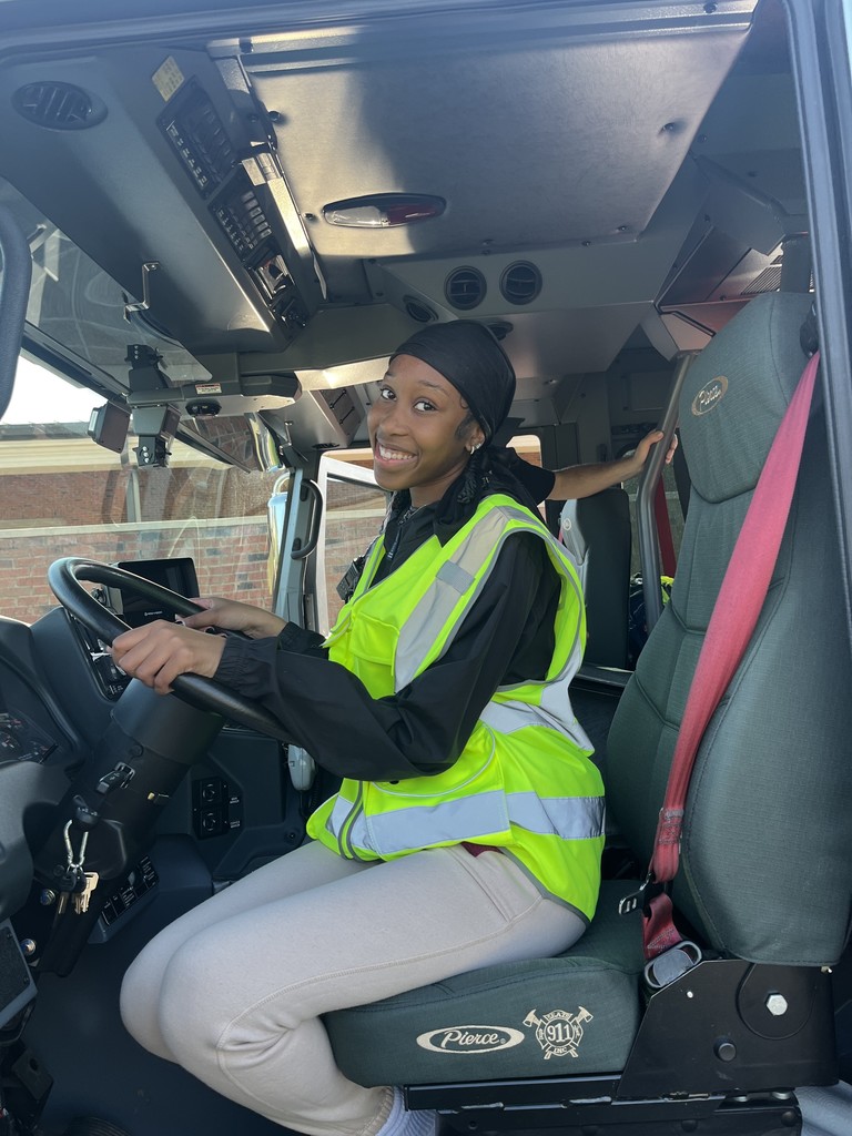 A smiling student wearing a neon yellow safety vest sits in the driver’s seat of a fire truck, holding the steering wheel.