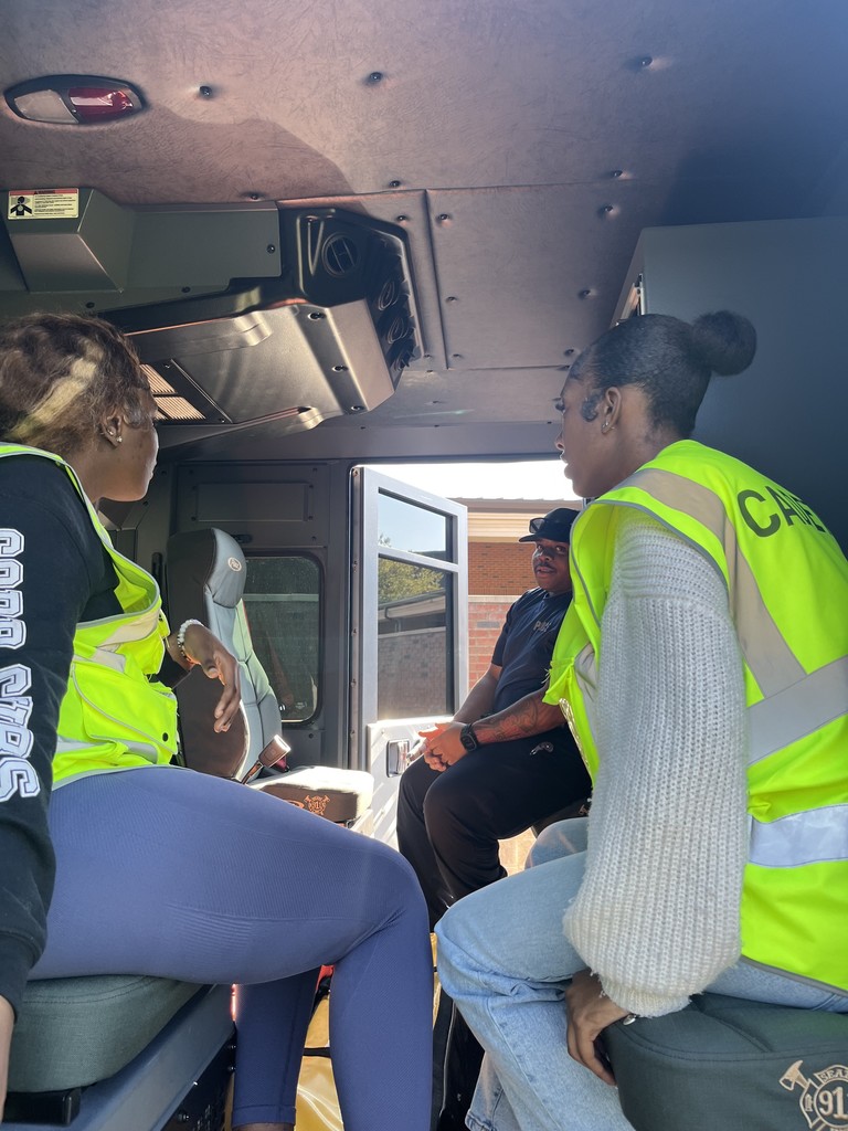 Two students in safety vests sit inside the cab of the fire truck, listening to a Bainbridge Public Safety officer who is seated near the open door.