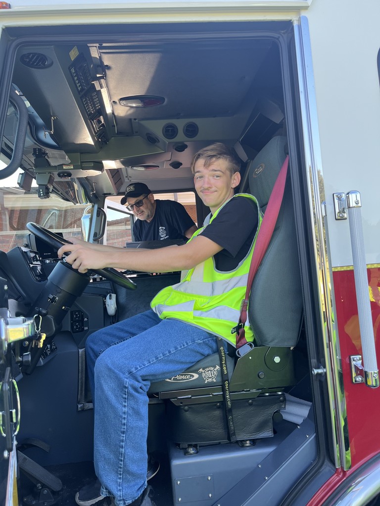 A student wearing a safety vest sits in the fire truck driver’s seat smiling, with a BPS firefighter standing nearby, guiding him.