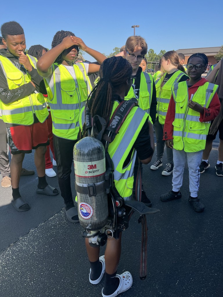 A student wearing a safety vest tries on a firefighter’s breathing apparatus while a BPS firefighter instructs him, as classmates watch in the background.