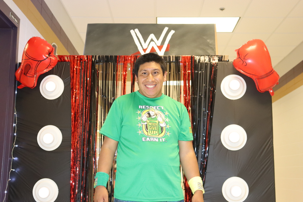 A smiling student wearing a green John Cena “Never Give Up” T-shirt stands in front of a WWE-themed backdrop decorated with red and black streamers, inflatable boxing gloves, and lights.