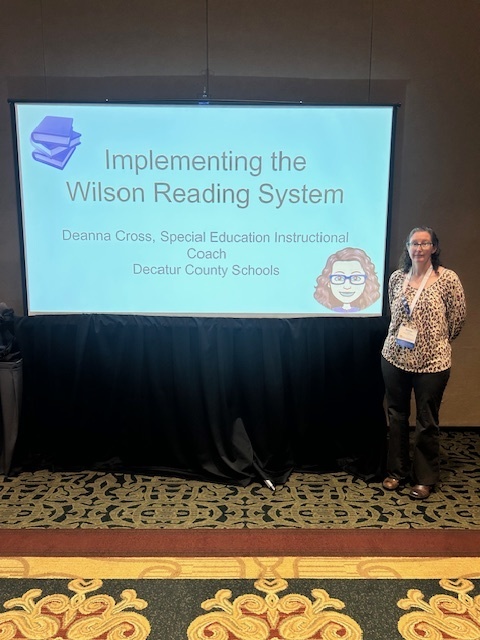 Deanna Cross, Special Education Instructional Coach at Decatur County Schools, standing beside a presentation slide titled 'Implementing the Wilson Reading System.'