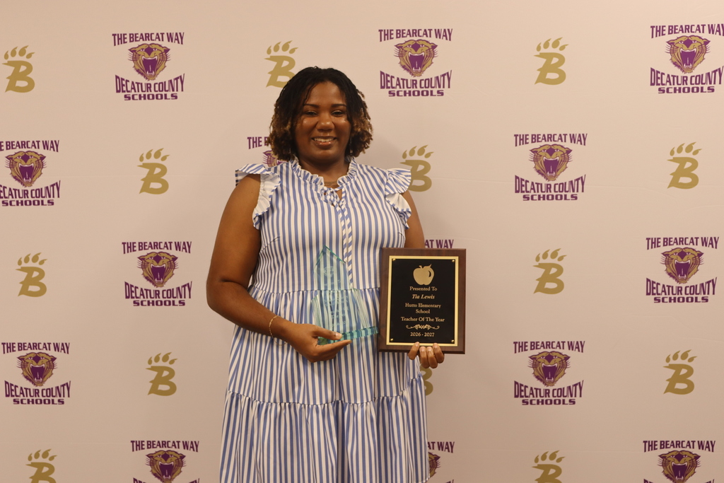A woman smiles while holding a glass award and a plaque that reads “Presented to Tia Lewis, Hutto Elementary School, Teacher of the Year, 2026–2027” in front of a Decatur County Schools backdrop.