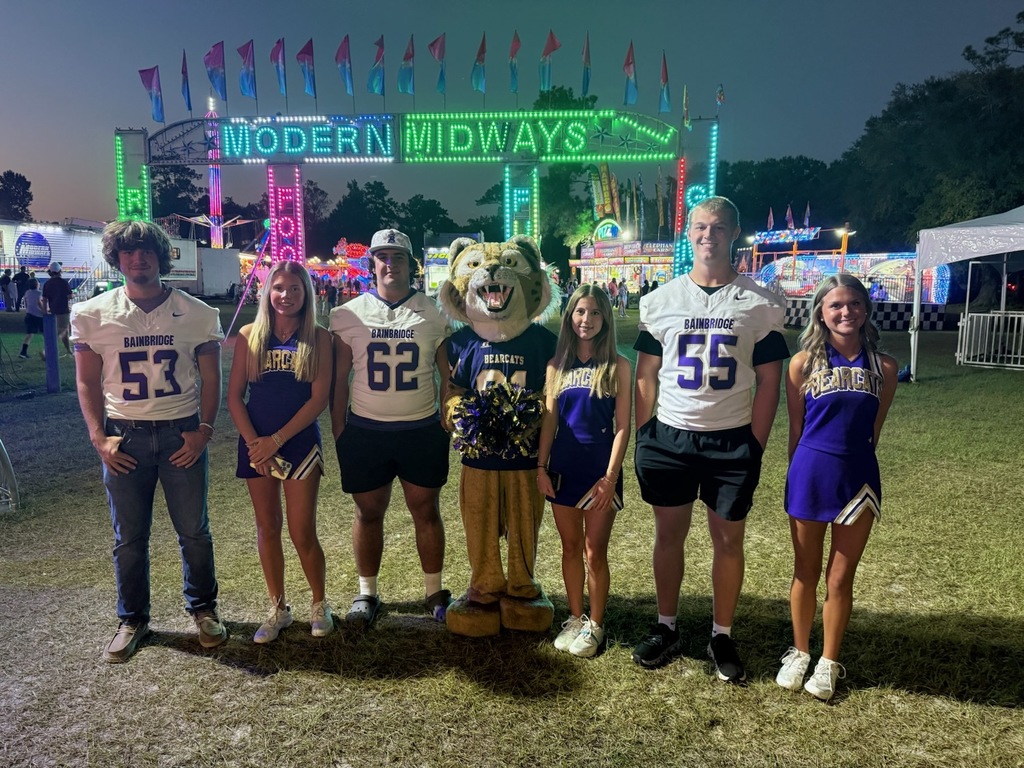 group of cheerleaders, football players and mascot stand in front of the fair entrance at dusk. the fair lights are glowing in the background