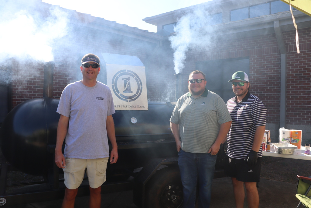three men standing in front of a grill with a metal sign on top that has the First National Bank logo lazered out of the metal