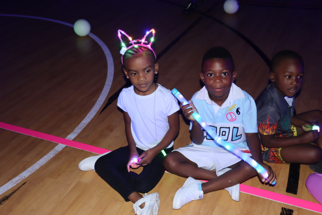 two primary students sit on a gym floor wearing rave headbands with a glow stick in hand