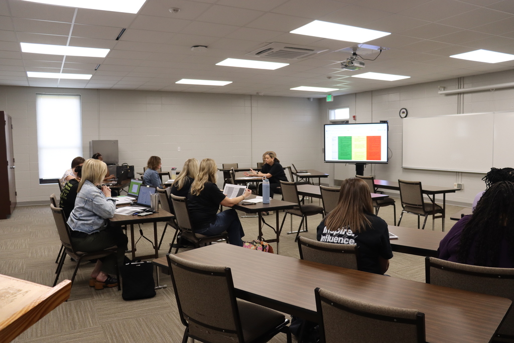 Educators seated in a classroom setting, listening to a presentation with a digital screen at the front.