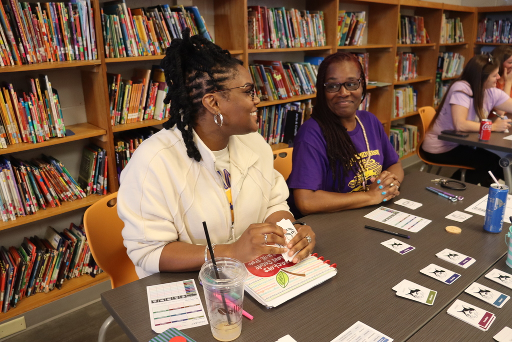 Two teachers smiling and talking at a table in a library during a collaborative activity.