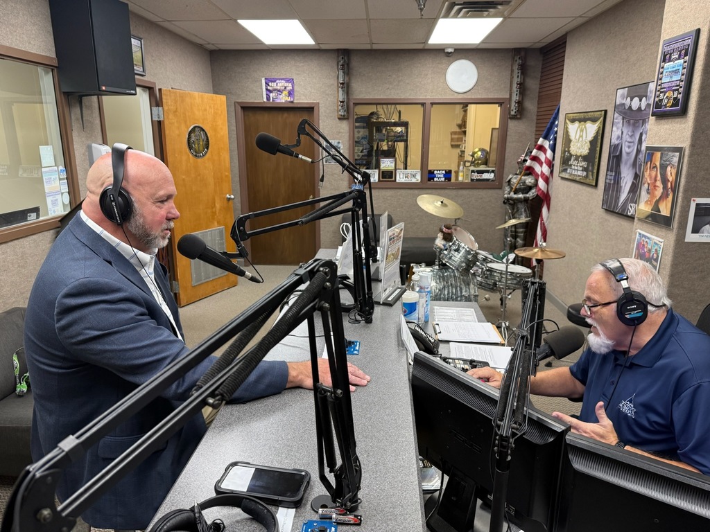 A man in a suit and another man in a polo shirt are speaking into microphones during a radio interview inside a studio with recording equipment and an American flag in the background.