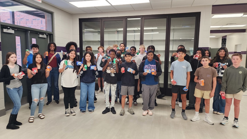 a group of students gather excitedly for a picture holding a snack and soda in a school hallway