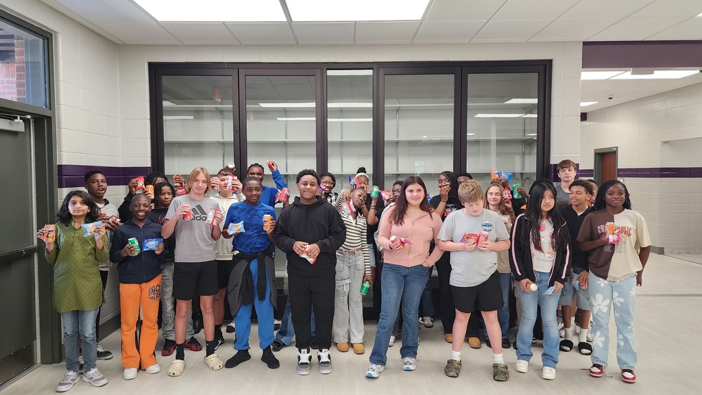 a group of students gather excitedly for a picture holding a snack and soda in a school hallway