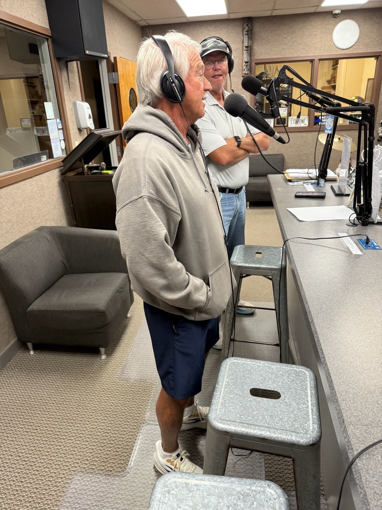 Two men wearing headphones speak into microphones in a radio studio. One man stands at the counter in a gray hoodie, navy shorts, and white sneakers, while the other stands behind him in a light gray polo shirt and cap.