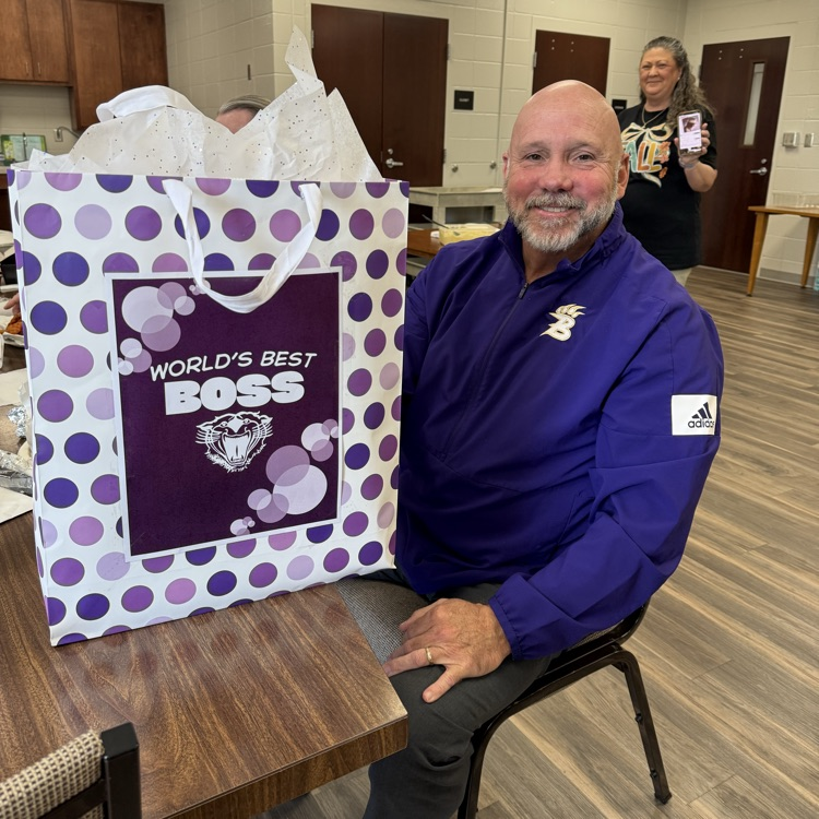 Dr. Boyd English smiles while sitting at a table holding a large purple and white gift bag that reads “World’s Best Boss” with a Bearcat logo on the front.