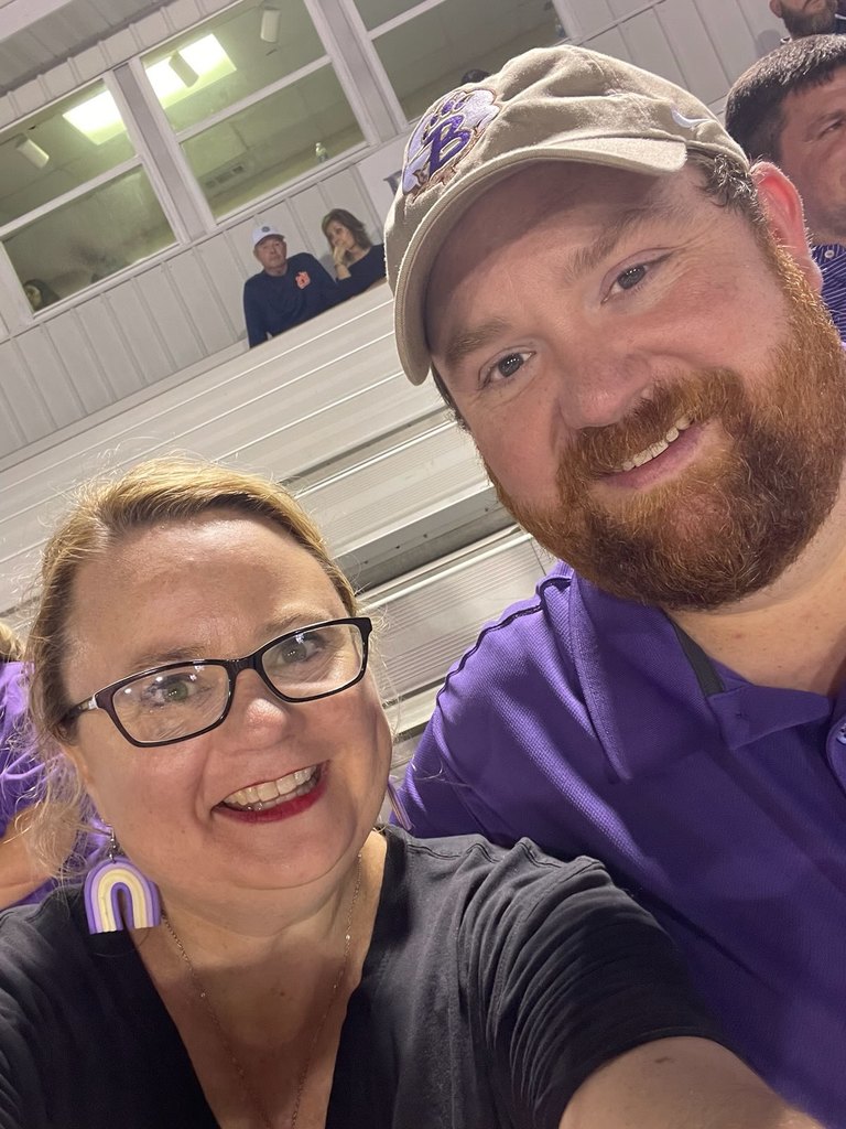 A smiling couple, both wearing Bainbridge Bearcat purple, sit together in the bleachers at a BHS football game. The man wears a Bearcat cap, and the woman wears rainbow earrings and glasses.