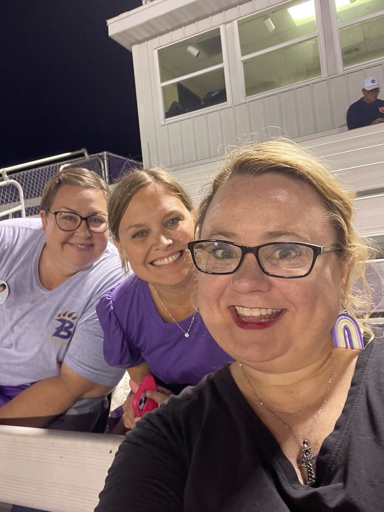 Three smiling teachers sit together in the bleachers at a Bainbridge High School football game. They are wearing purple and gray Bearcat gear, showing school spirit under the stadium lights.