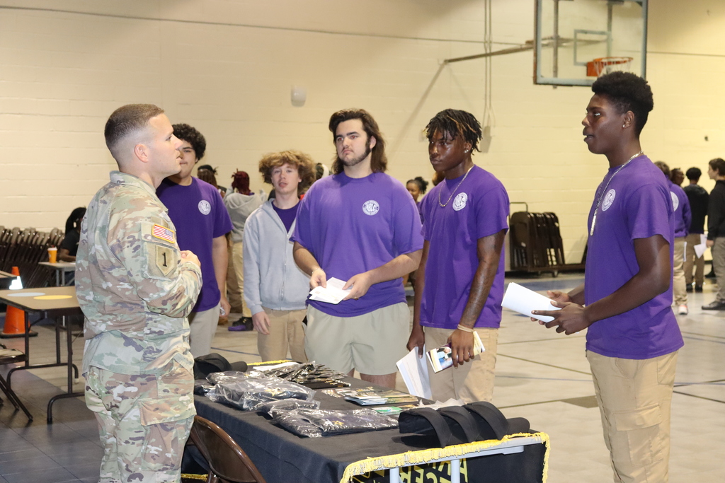 A U.S. Army recruiter speaks with a group of students wearing purple shirts at a career fair table.