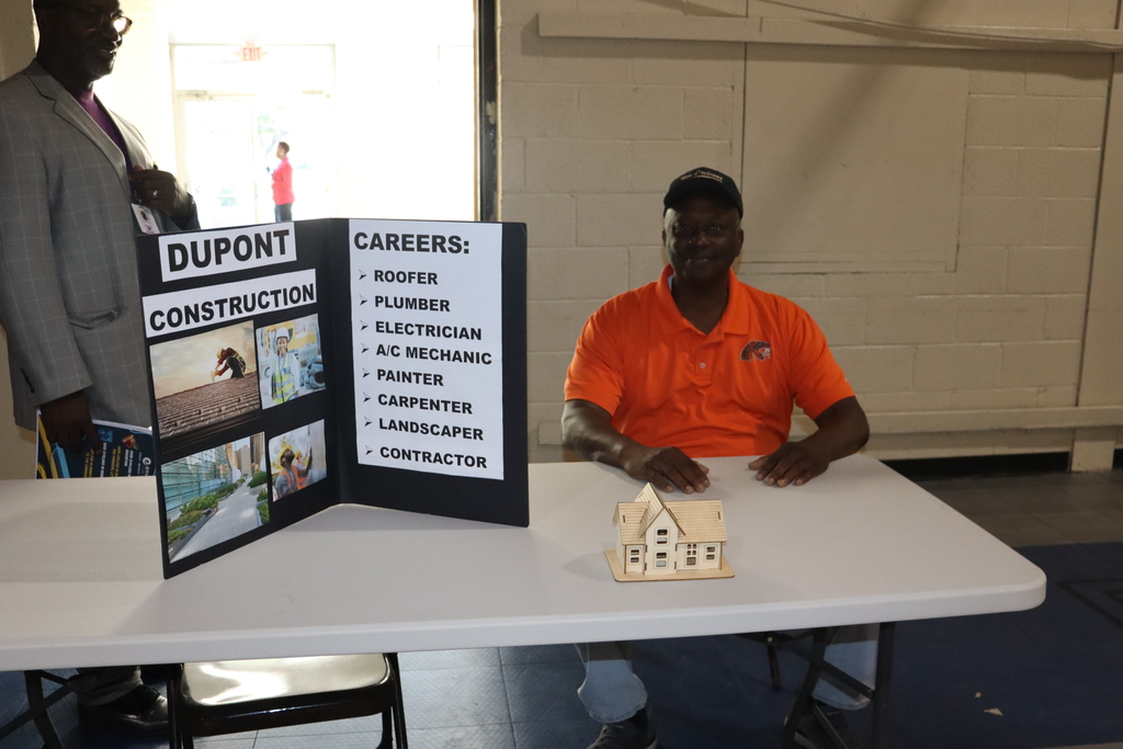A man at a Dupont Construction booth displays a poster listing career opportunities such as roofer, plumber, and electrician.