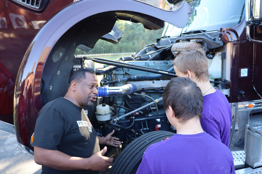 A trucking professional explains engine components to two students beside a large truck.