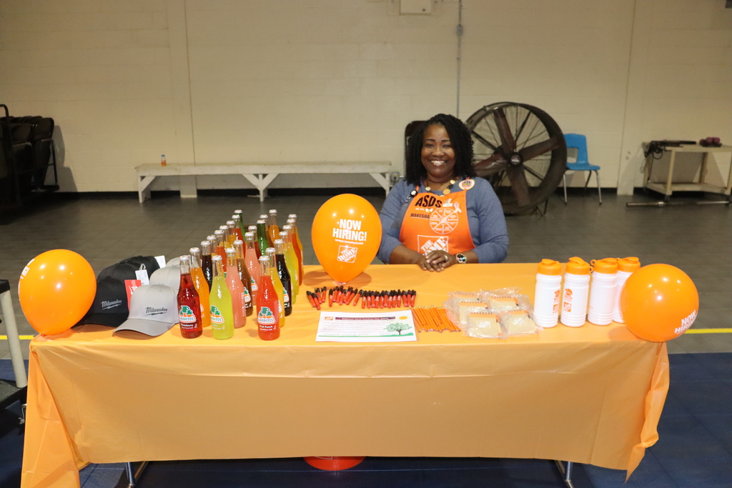 A Home Depot representative smiles at a career fair booth decorated with orange balloons, bottled drinks, and giveaway items.