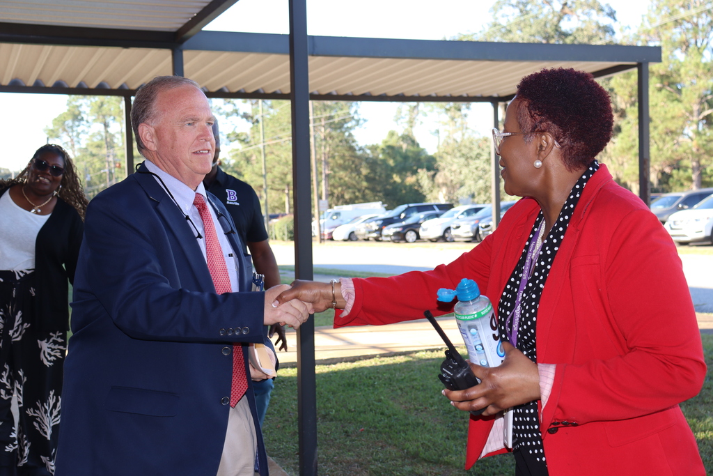 A man in a navy suit shakes hands with a woman in a red blazer outside the school building, greeting attendees.