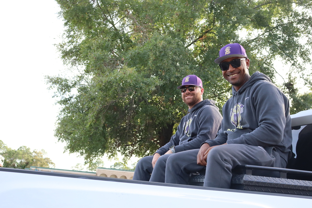 Two Bainbridge coaches in purple hats and hoodies ride on the back of a truck, smiling during the parade.