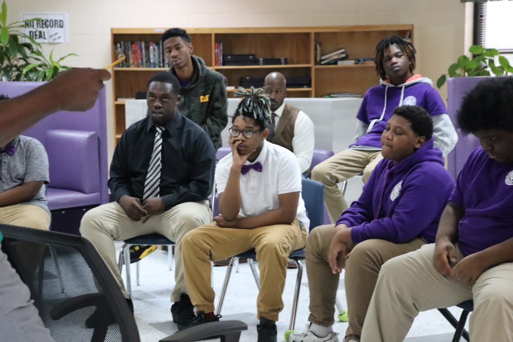 A group of students and mentors sit attentively in a library setting during a Tie Tuesday Gentlemen’s Club meeting.