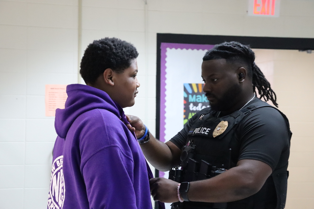 A police officer helps a student in a purple hoodie adjust his tie in a school hallway.