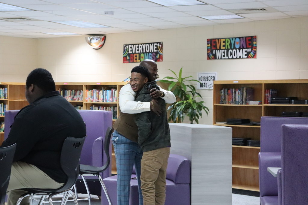 Two men share a warm hug in a school library setting, surrounded by bookshelves and signs reading “Everyone Belongs” and “Everyone Is Welcome.”