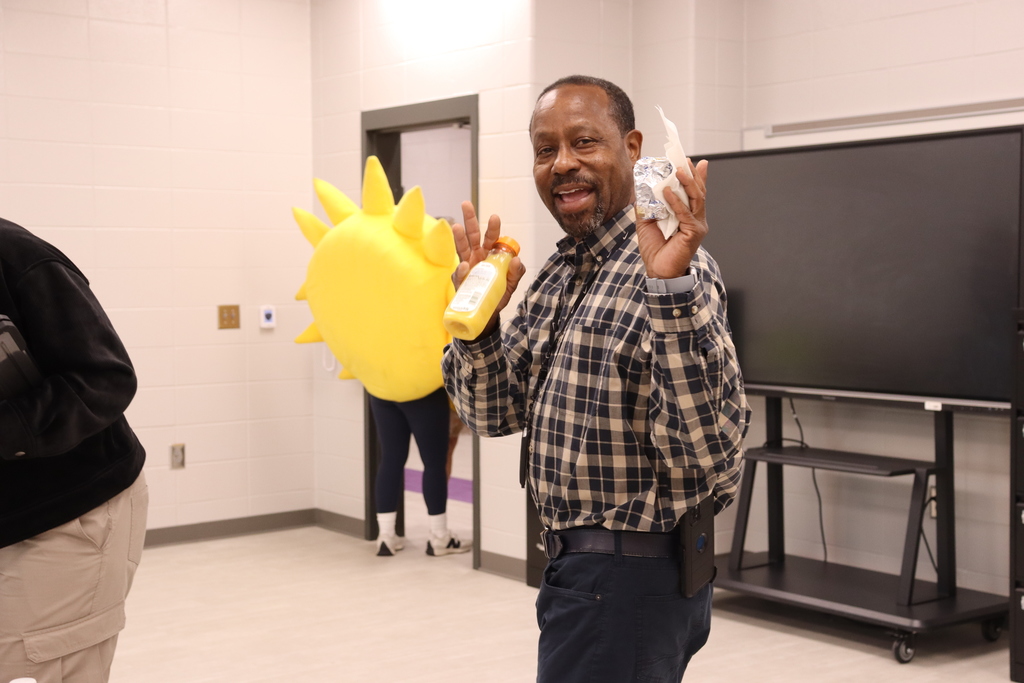 A BMS staff member smiles while holding a biscuit and orange juice, with Sunny the SunStop mascot in the background.