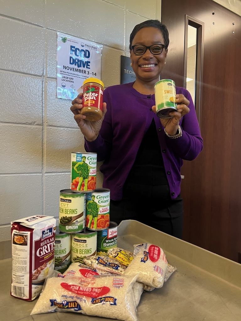 A woman wearing a purple sweater smiles while holding a jar of peanut butter and a can of corn. In front of her is a table filled with donated food items, including canned vegetables, rice, and grits, for a community food drive.