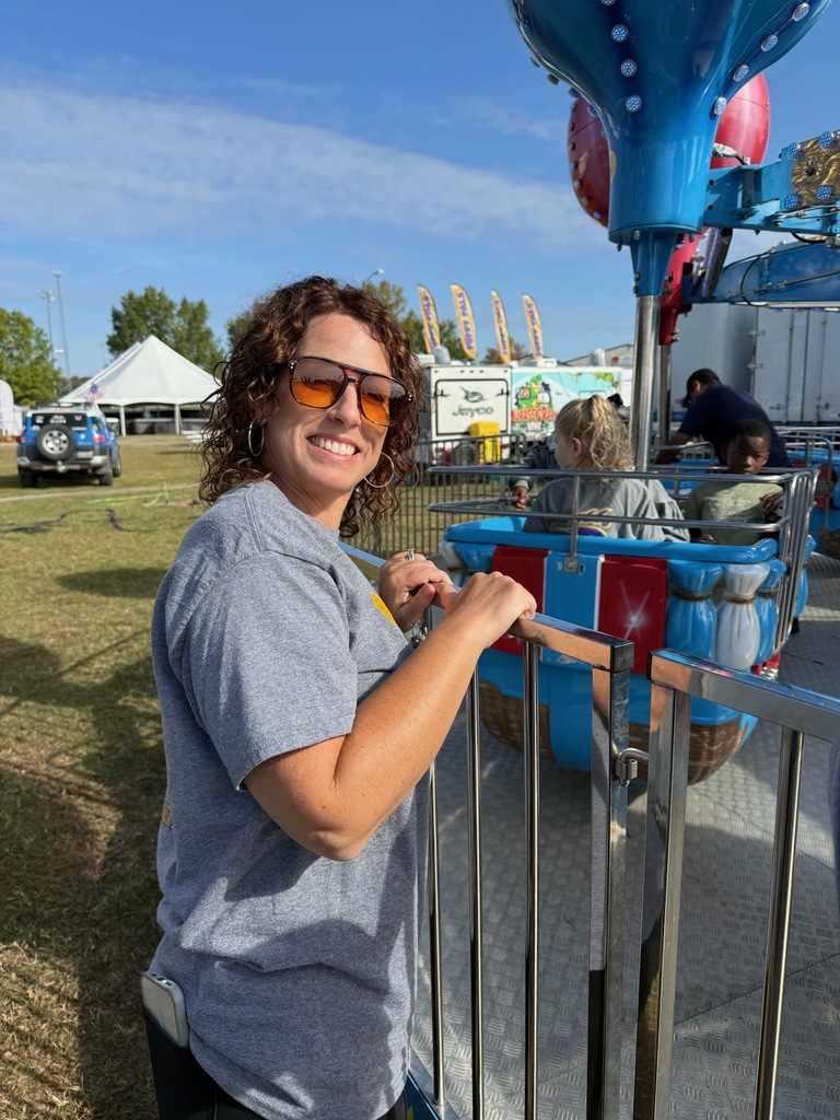 Woman smiling by a carnival ride on a sunny day, wearing sunglasses and a gray shirt.