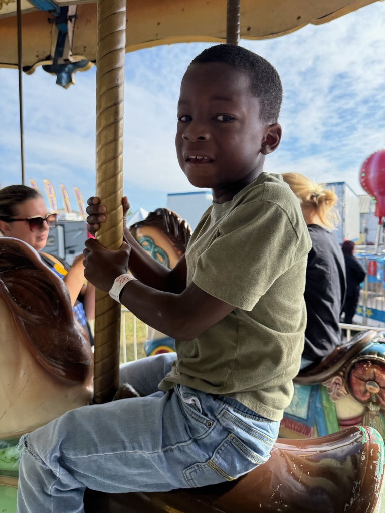 Young boy rides a carousel horse at a fair on a bright, sunny day.