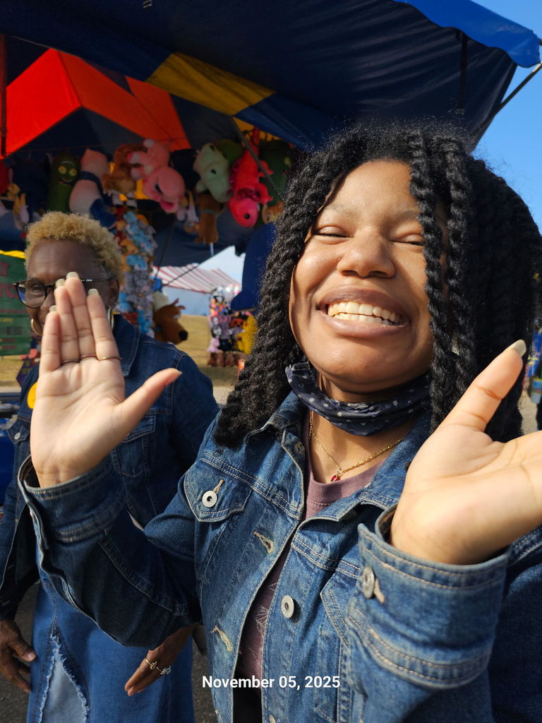 Smiling girl in a denim jacket waves joyfully at a fair, with colorful stuffed animals in the background.