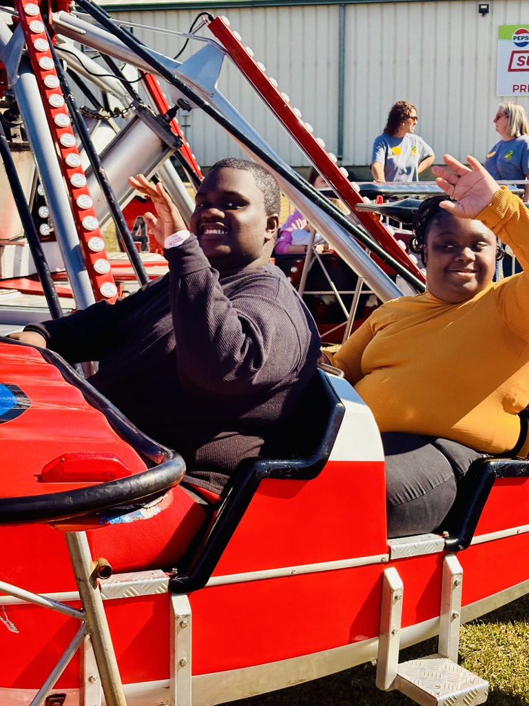 Two people smiling and waving while seated on a red and white amusement park ride, enjoying a sunny day outdoors.