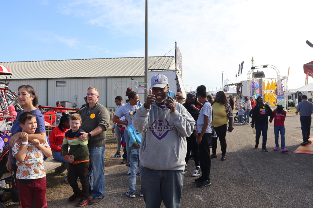 Man smiling and giving a thumbs-up while talking on the phone at a busy fairground.