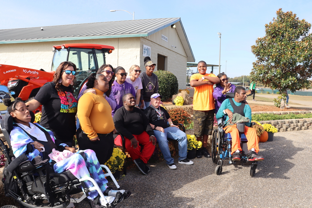 A cheerful group poses together outdoors in front of flowers and a tractor on a sunny day.