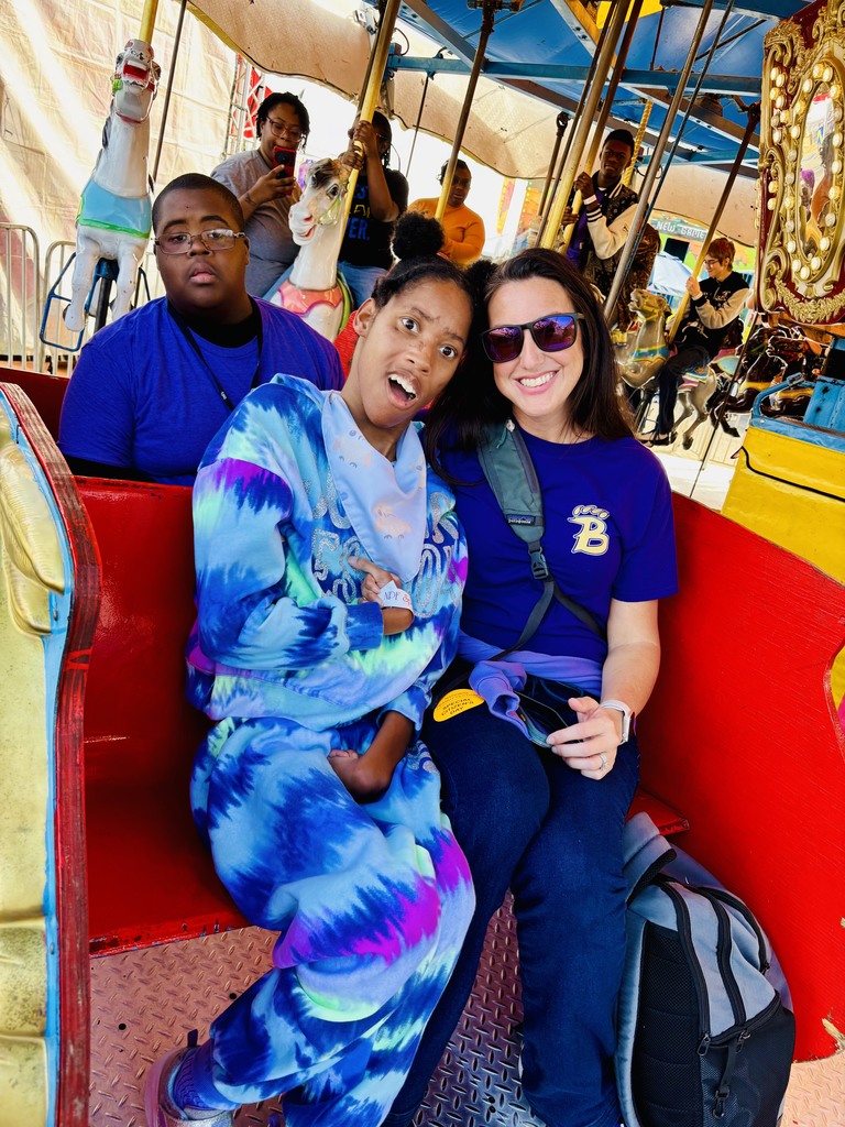 Smiling group enjoys a carousel ride, with one person wearing a colorful tie-dye outfit and another in a purple shirt with a “B” logo.