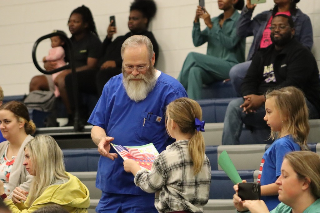A kindergarten student hands a colorful Veterans Day card to an older man in blue scrubs during the JWP Veterans Day program.