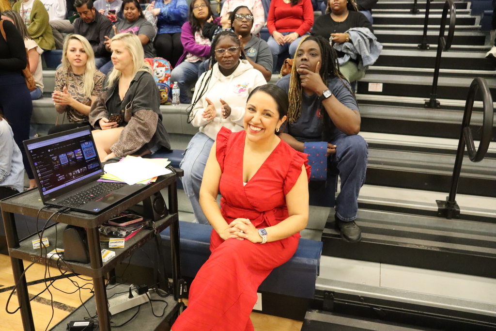 A smiling woman in a red outfit sits near sound equipment while the audience applauds behind her.