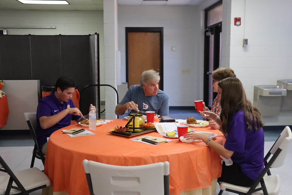 Students and community members sit together at a round table eating lunch during the Community Task Force event. Plates of food and decorative centerpieces are on the table.