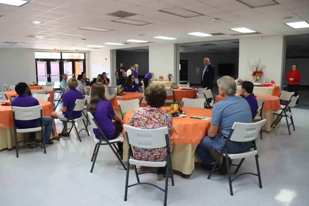 A wide view of the Community Task Force meeting room shows students and community members seated at several round tables, listening to a speaker standing near the side of the room.
