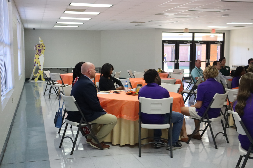 Community Task Force participants sit at round tables covered with orange tablecloths inside a large meeting room. Principal Bryant sits at one table alongside students and community members.