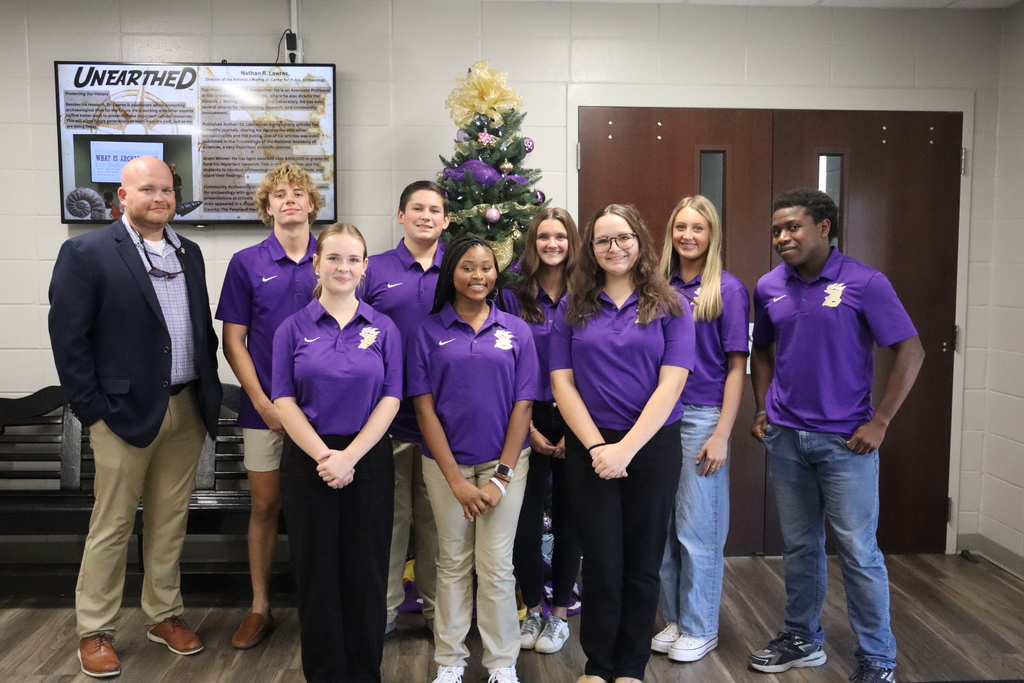 A group of Bainbridge High School Bearcat Ambassadors wearing purple polos stand together indoors with Principal Bryant, who is on the far left. A decorated Christmas tree and a display screen are visible in the background.