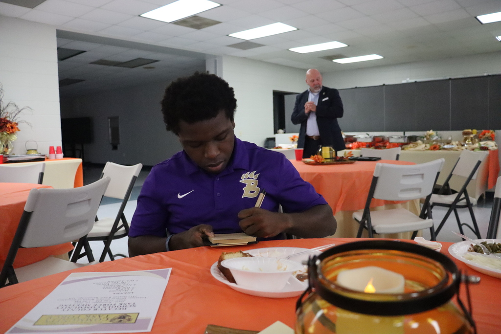 A Bearcat Ambassador student sits at a table writing in a small notebook after the meal. In the background, Superintendent Dr. English stands near the buffet table.