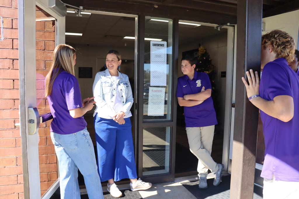 Bearcat Ambassador students stand at the entrance of a building, talking with an adult wearing a white jacket and blue skirt. Two students lean casually near the doorframe while chatting.
