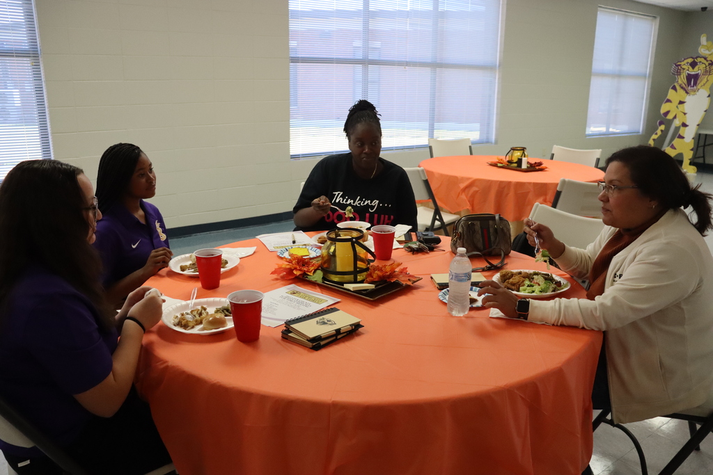 A group of students and community members share a meal at a round table decorated with fall-themed items. They are engaged in conversation while eating.